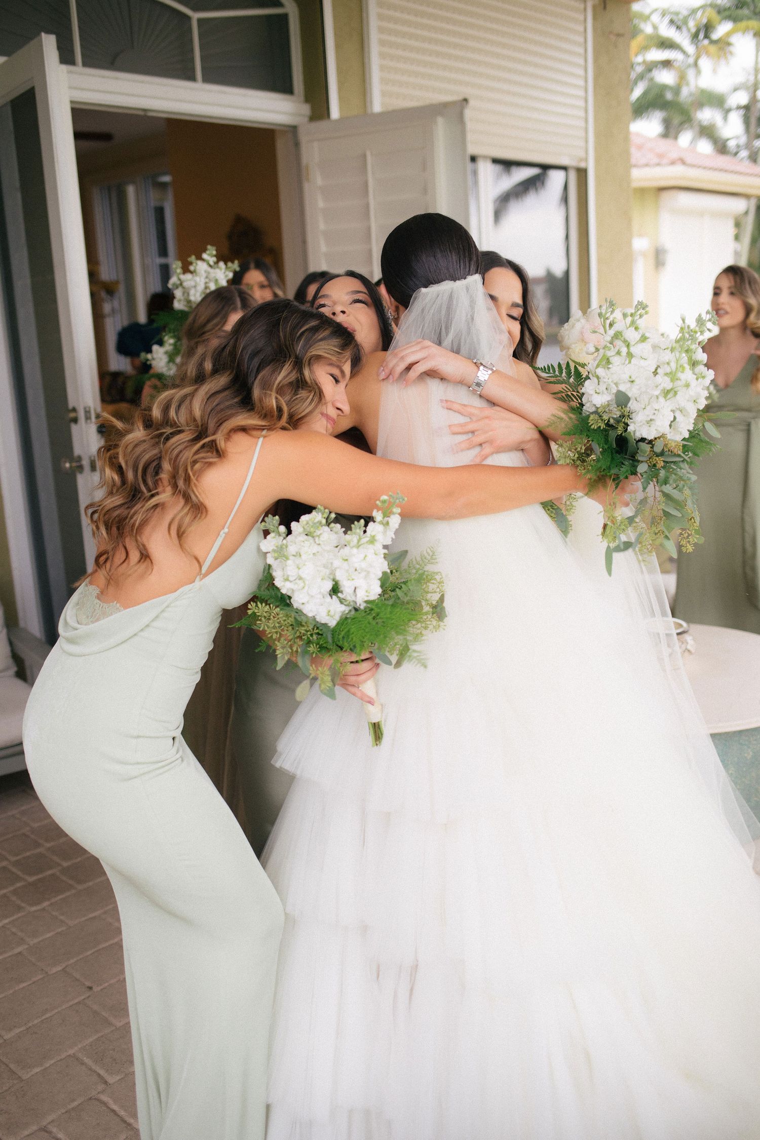 Bridesmaids in sage green dresses embrace the bride in a white gown and veil, holding white floral bouquets.