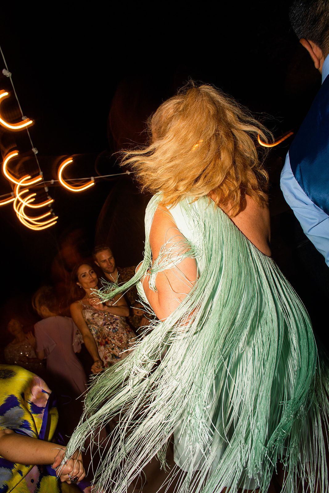 Person with blonde hair dancing energetically in a mint green dress with flowing fringe under dim party lights.