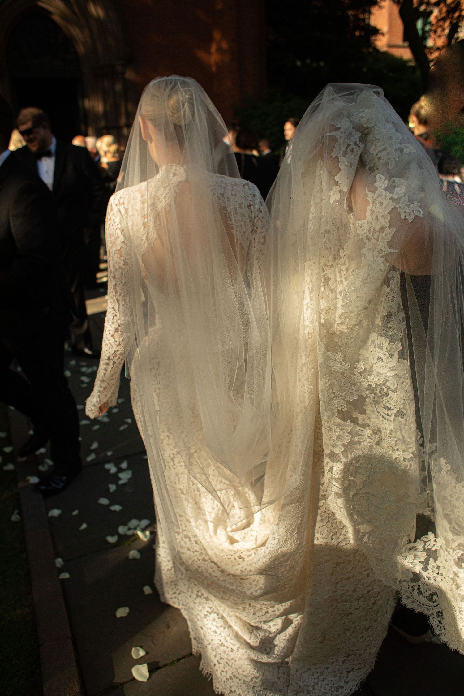 Two brides in elegant lace wedding gowns and veils walk together after their ceremony, rose petals scattered on the ground.