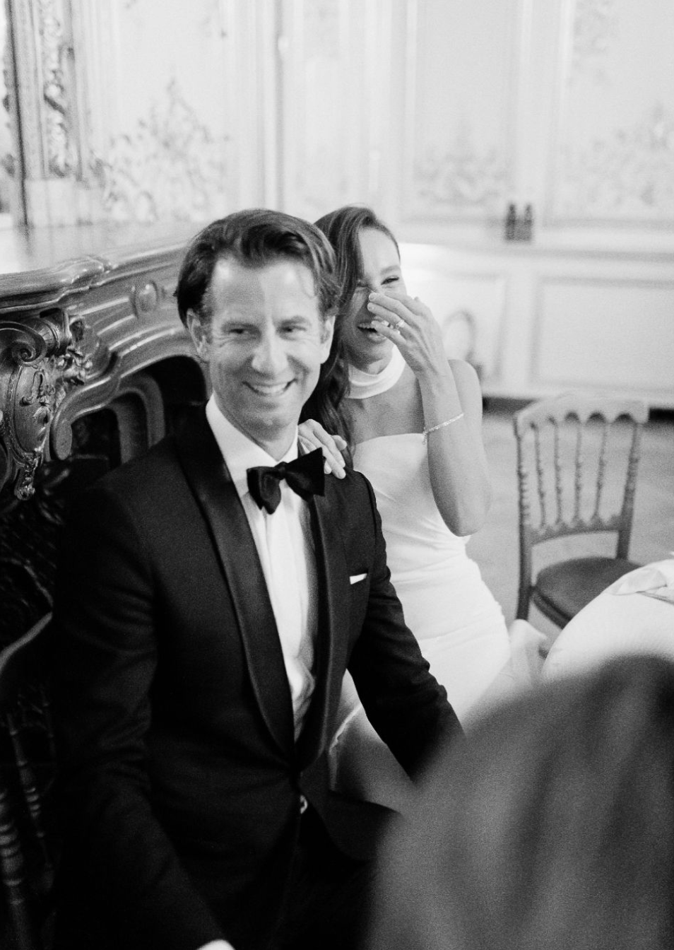 Black and white photo of a smiling groom in a tuxedo and bride laughing at their elegant wedding reception.