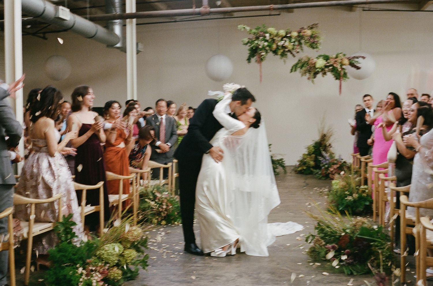 Bride and groom share a kiss in the aisle at an indoor wedding ceremony as seated guests cheer beneath hanging floral installations.