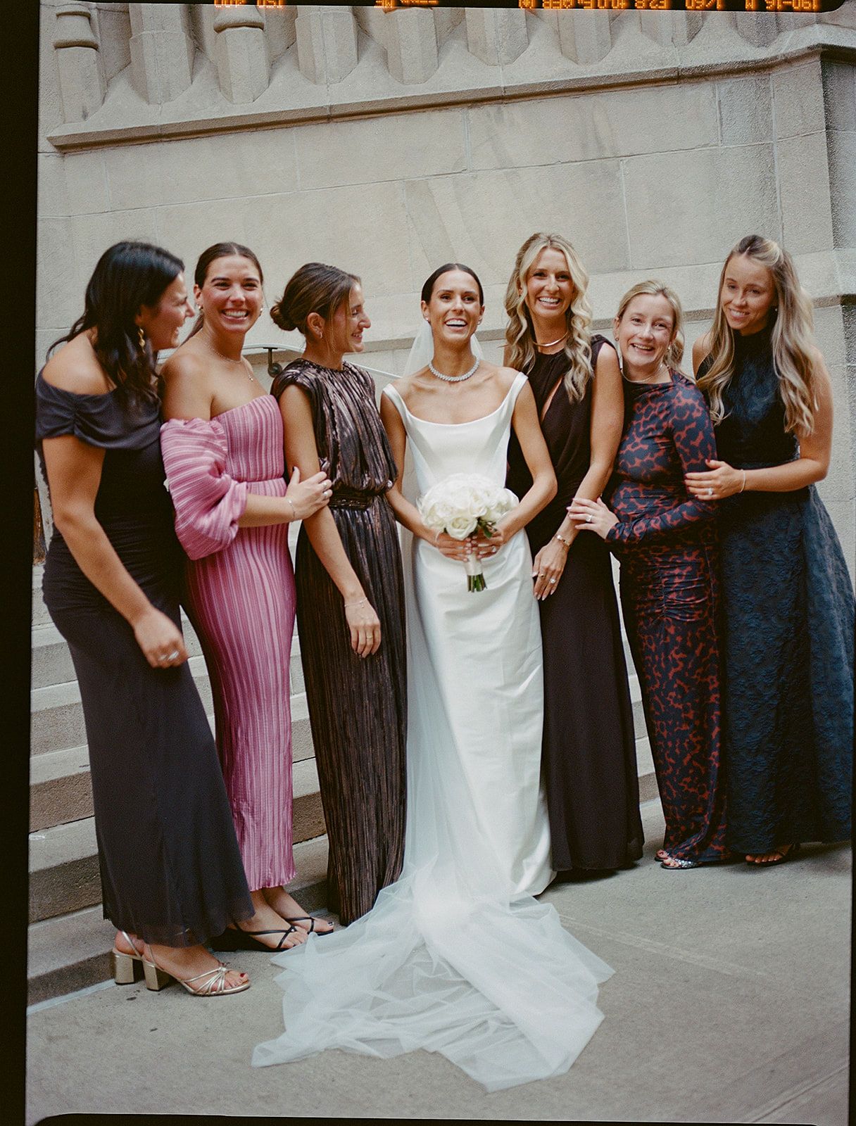 Wedding party in formal attire poses together against ornate wall, with bridesmaids in dark dresses and bride in white gown.