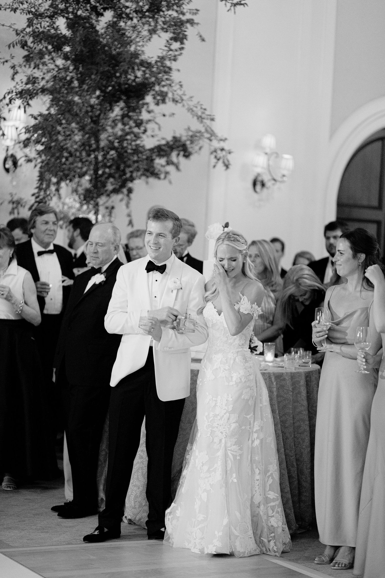 Black and white photo of a smiling bride and groom during wedding reception toasts, surrounded by elegantly dressed guests.
