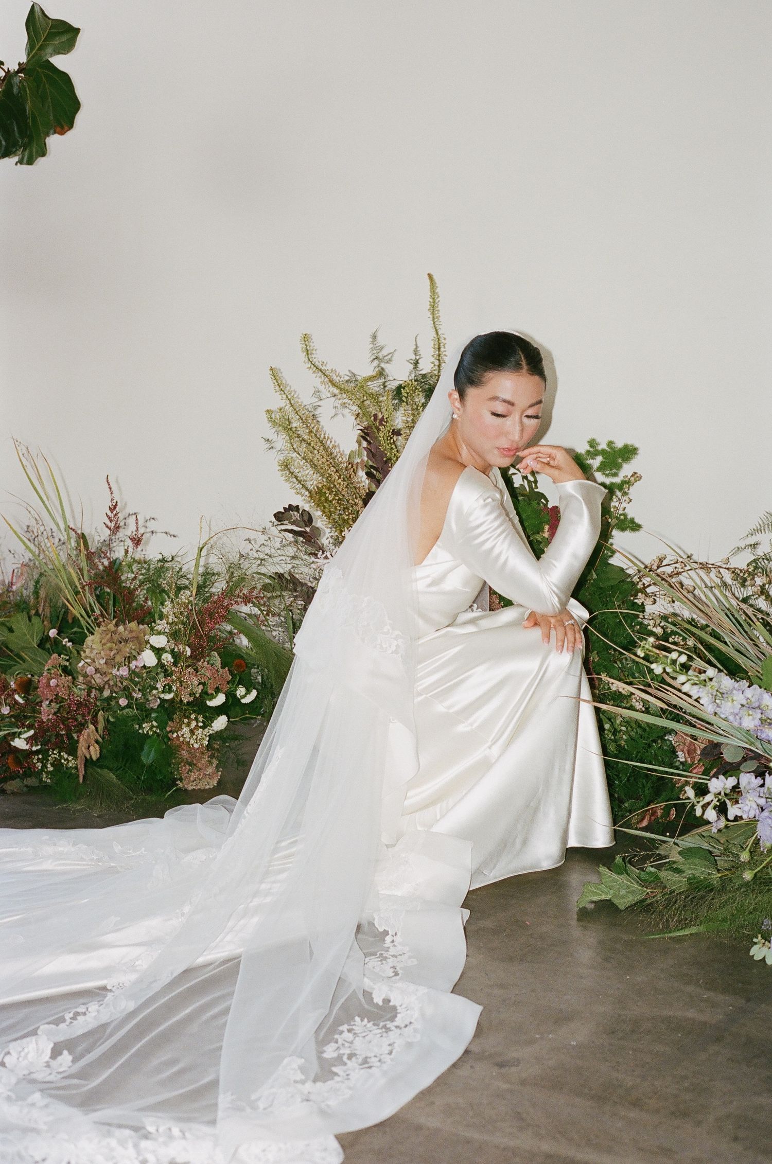 A bride in a flowing white silk gown and veil poses gracefully among floral arrangements and dried grasses.