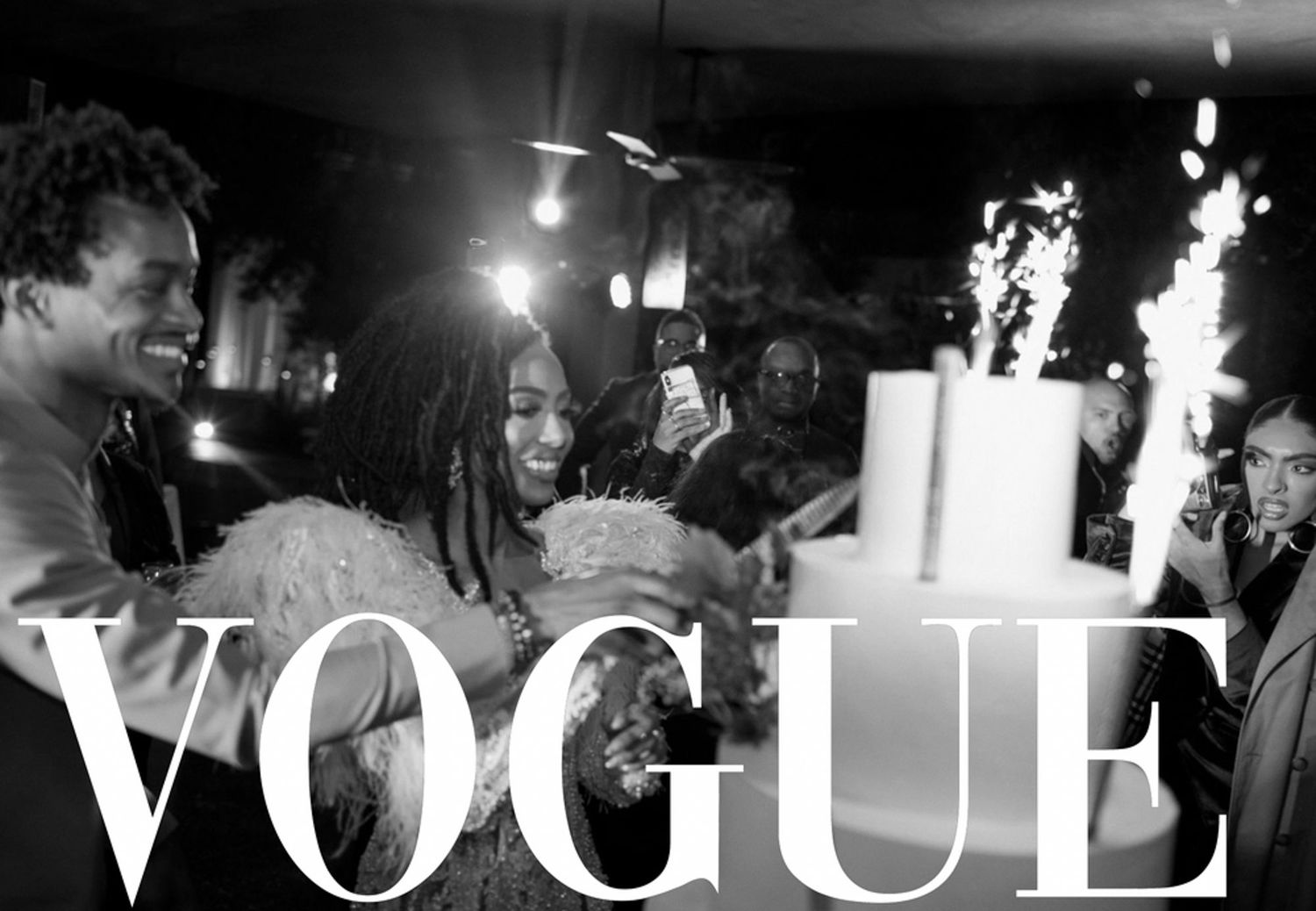 Black and white Vogue photo of people celebrating around a tiered cake with sparklers at an elegant party.