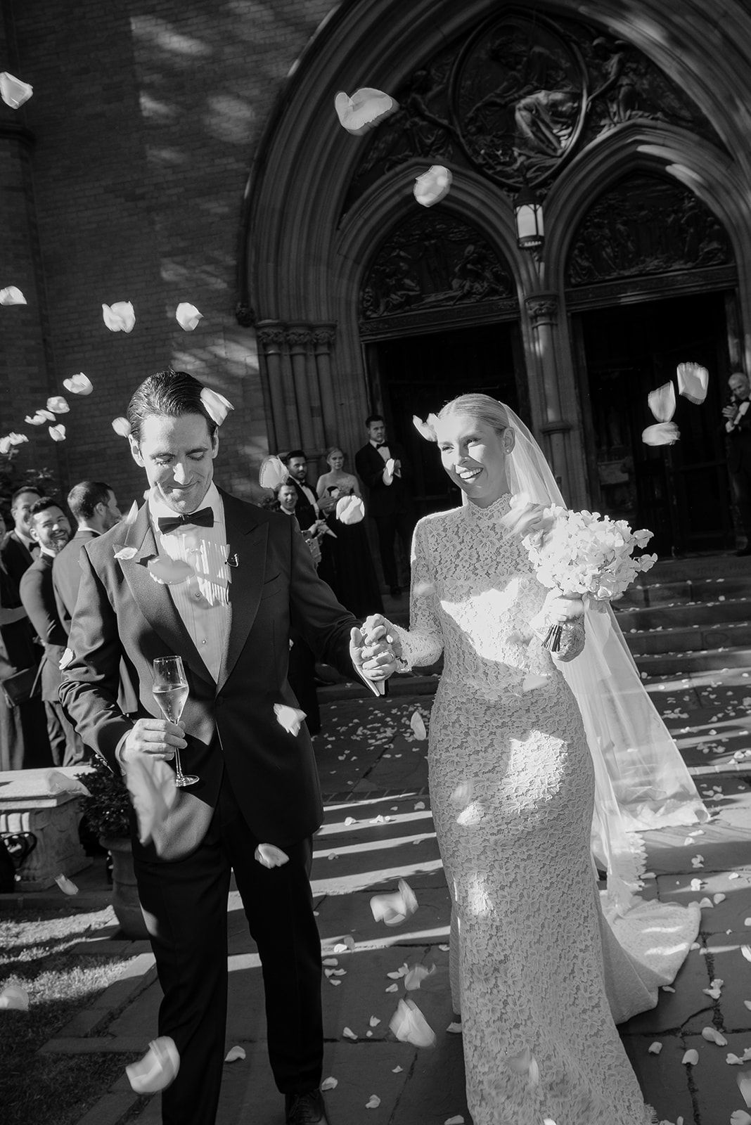Bride and groom hold hands and smile as petals shower them on the steps of a Gothic church after their wedding ceremony.
