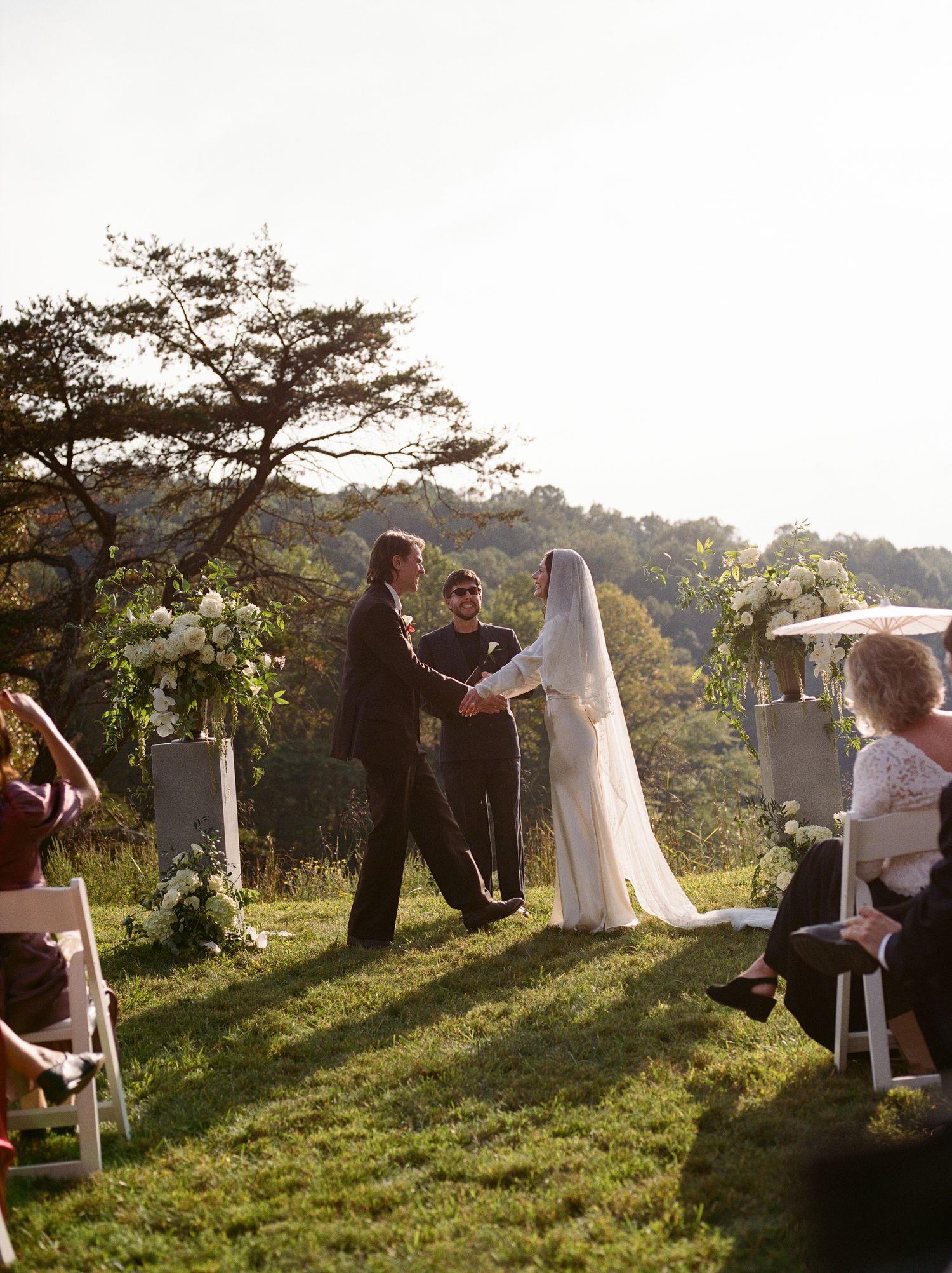 Bride in a long white veil and groom hold hands before an officiant during an outdoor wedding ceremony on a grassy hillside with guests.
