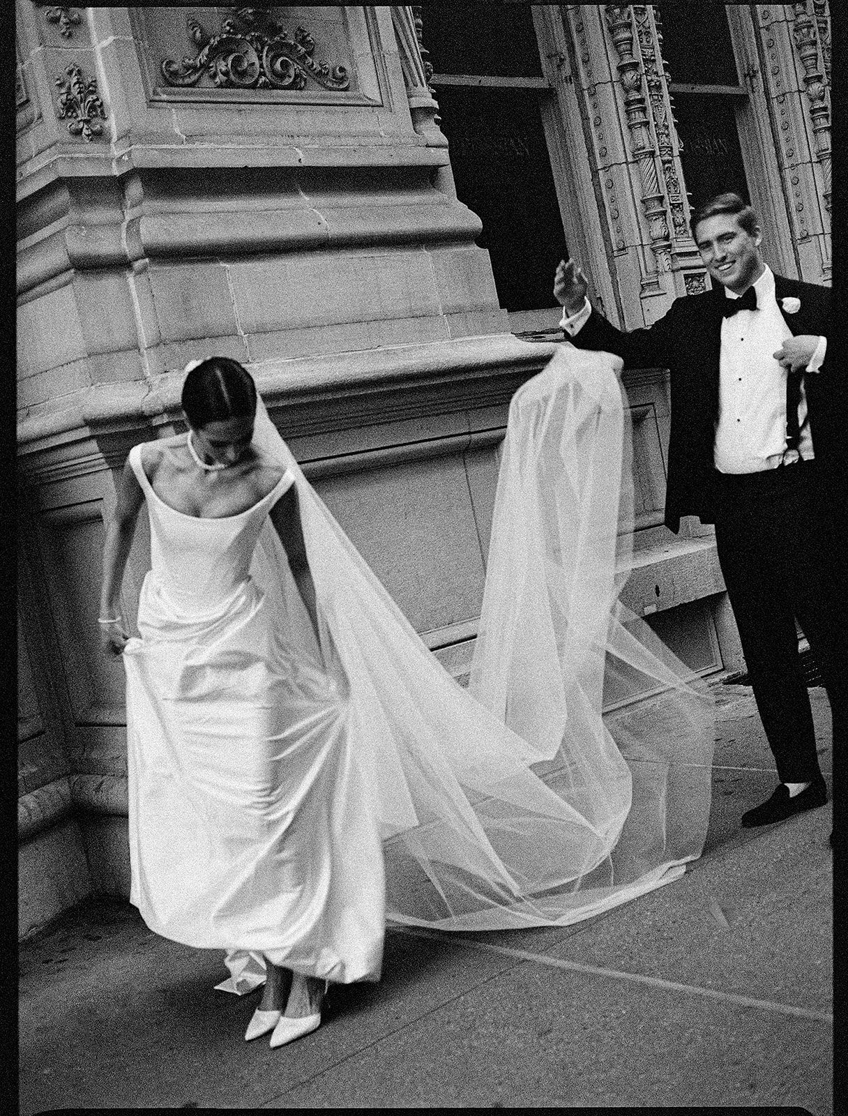 Black and white photo of a bride in a flowing gown and groom in tuxedo on elegant building steps.