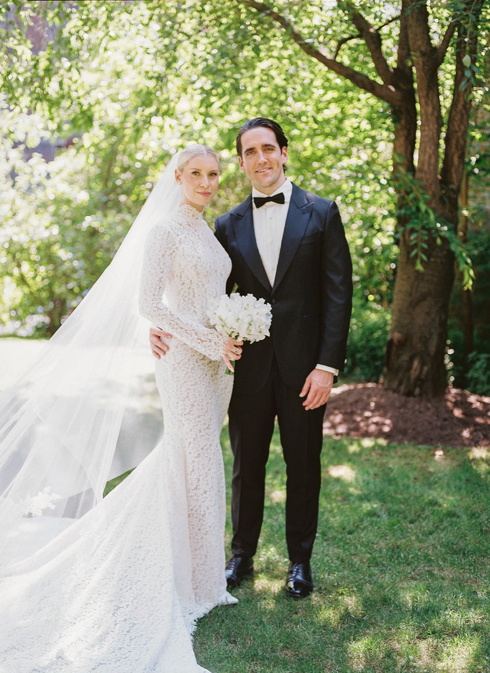 Elegant bride in long-sleeve lace gown with veil and groom in black tuxedo posing outdoors under green trees.