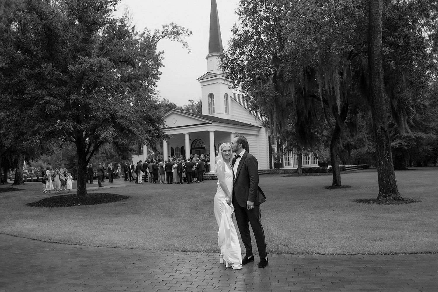 Black and white photo of a bride and groom kissing outside a white church with wedding guests gathered in the background.