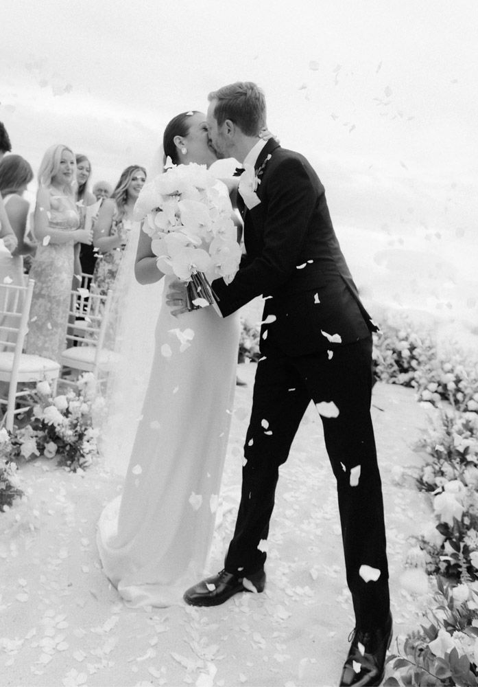Bride and groom share first kiss as flower petals fall during outdoor wedding ceremony.