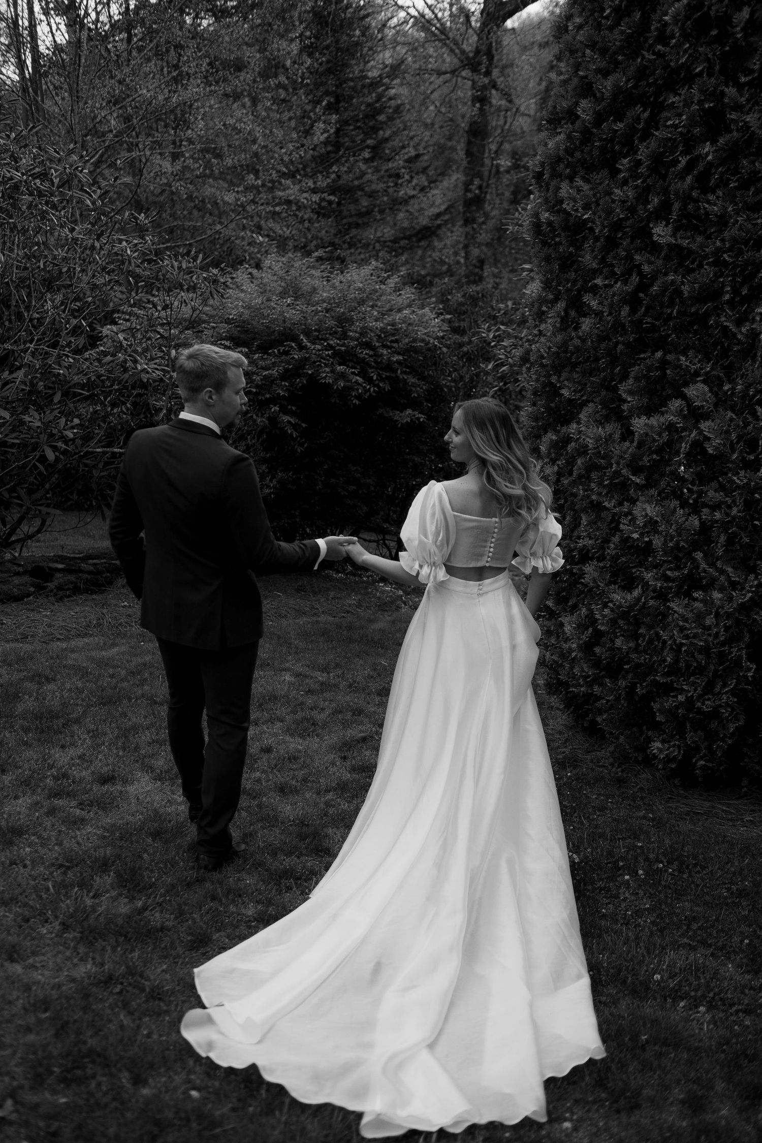 Black and white photo of a bride in a flowing white gown holding hands with a groom in a suit in a lush garden.