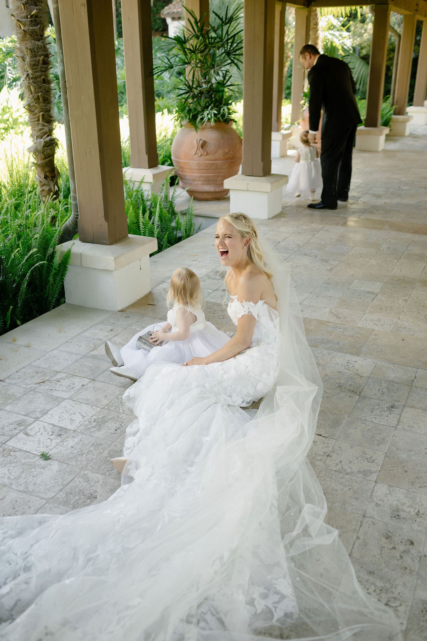 Bride in white gown laughs with flower girl on elegant marble terrace, groom visible in background.
