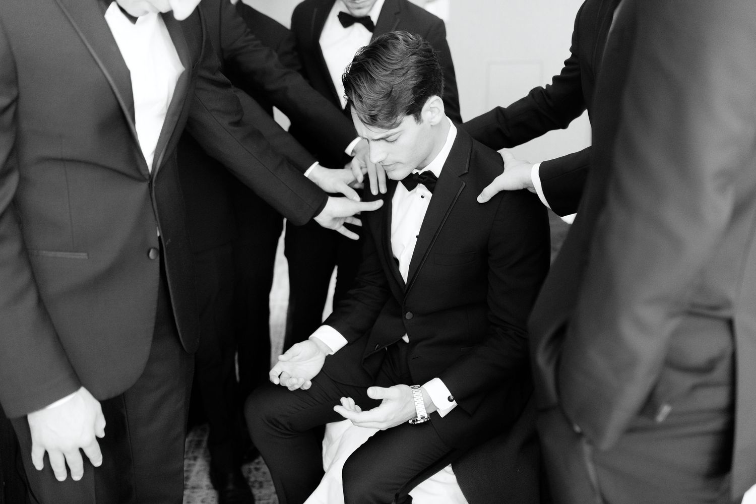 Groomsmen in black tuxedos gather around groom in candid black and white wedding photo.