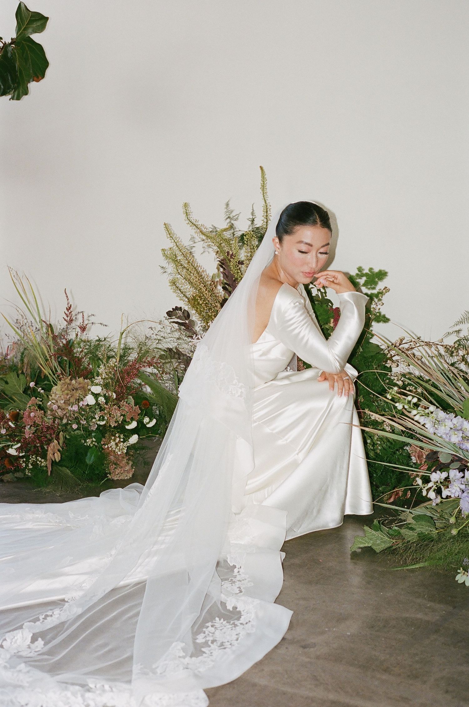Bride in elegant white satin gown with long lace-trimmed veil, posed gracefully among lush greenery and wildflowers.