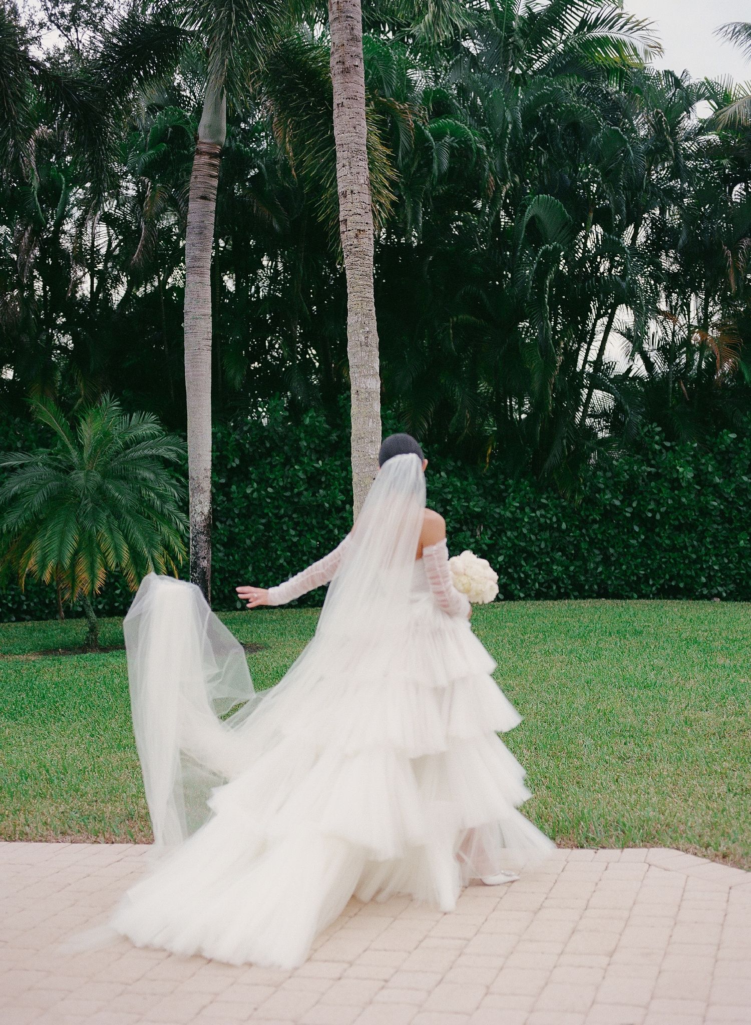 A flowing white wedding dress with delicate tulle layers billows gracefully on a stone pathway surrounded by tropical palm trees.