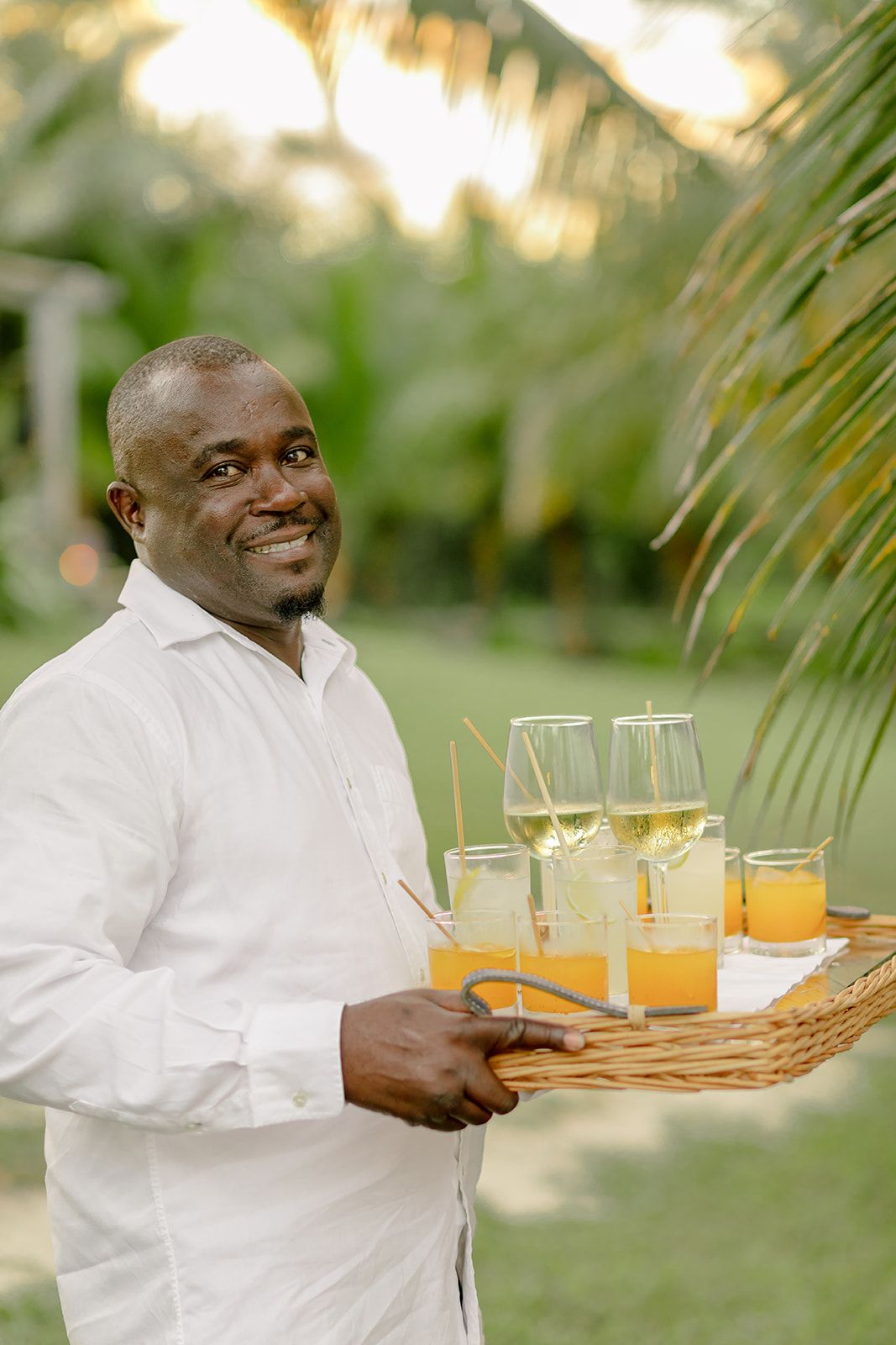 Server carrying wicker tray with champagne glasses and orange drinks on tropical resort patio.