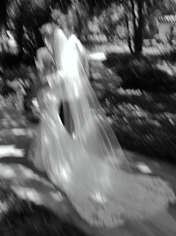 Blurry black and white photo of a bride in a flowing white wedding dress and veil outdoors.