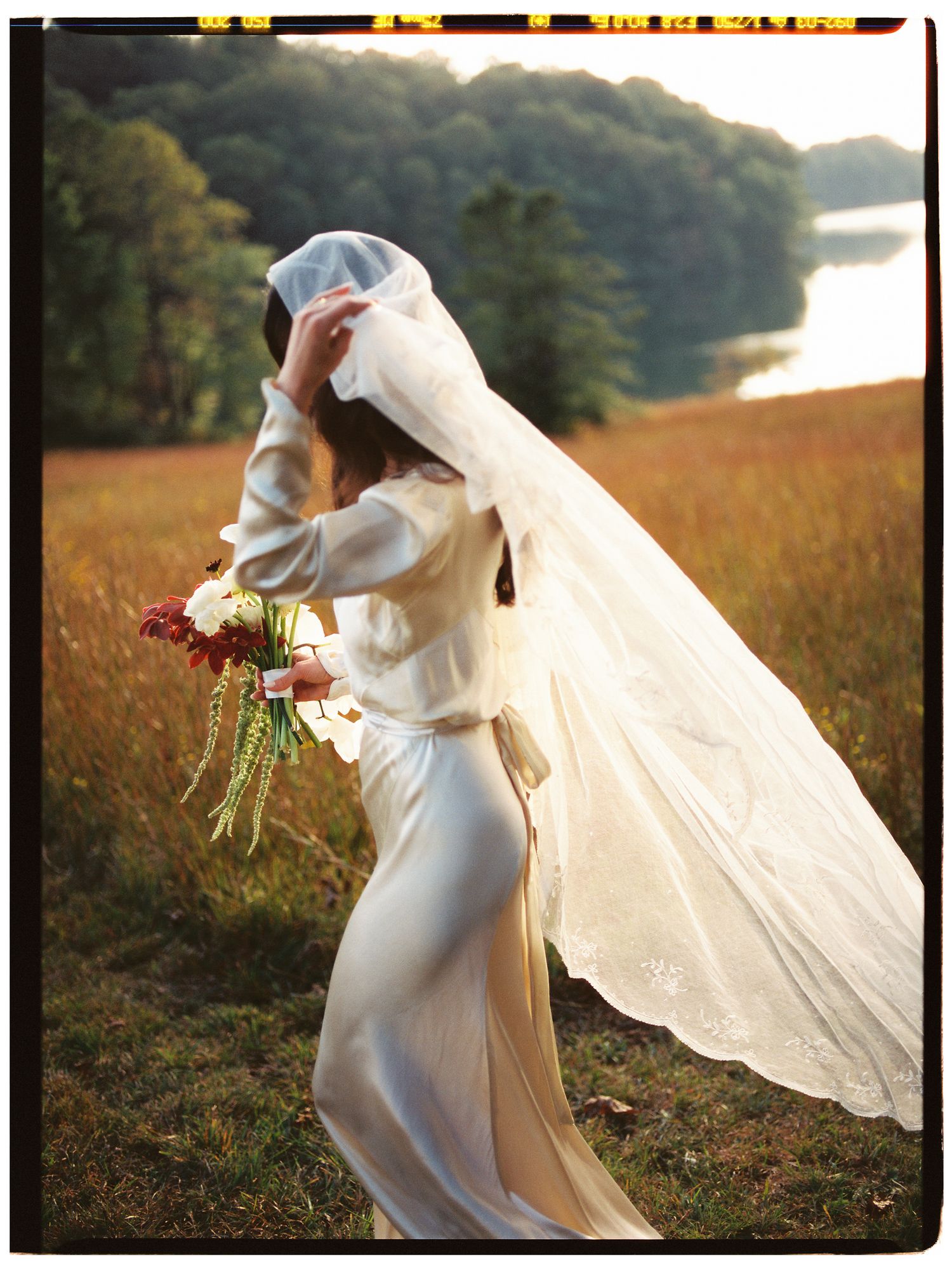 A flowing white wedding dress and veil catch the golden sunlight in a dreamy meadow at sunset.
