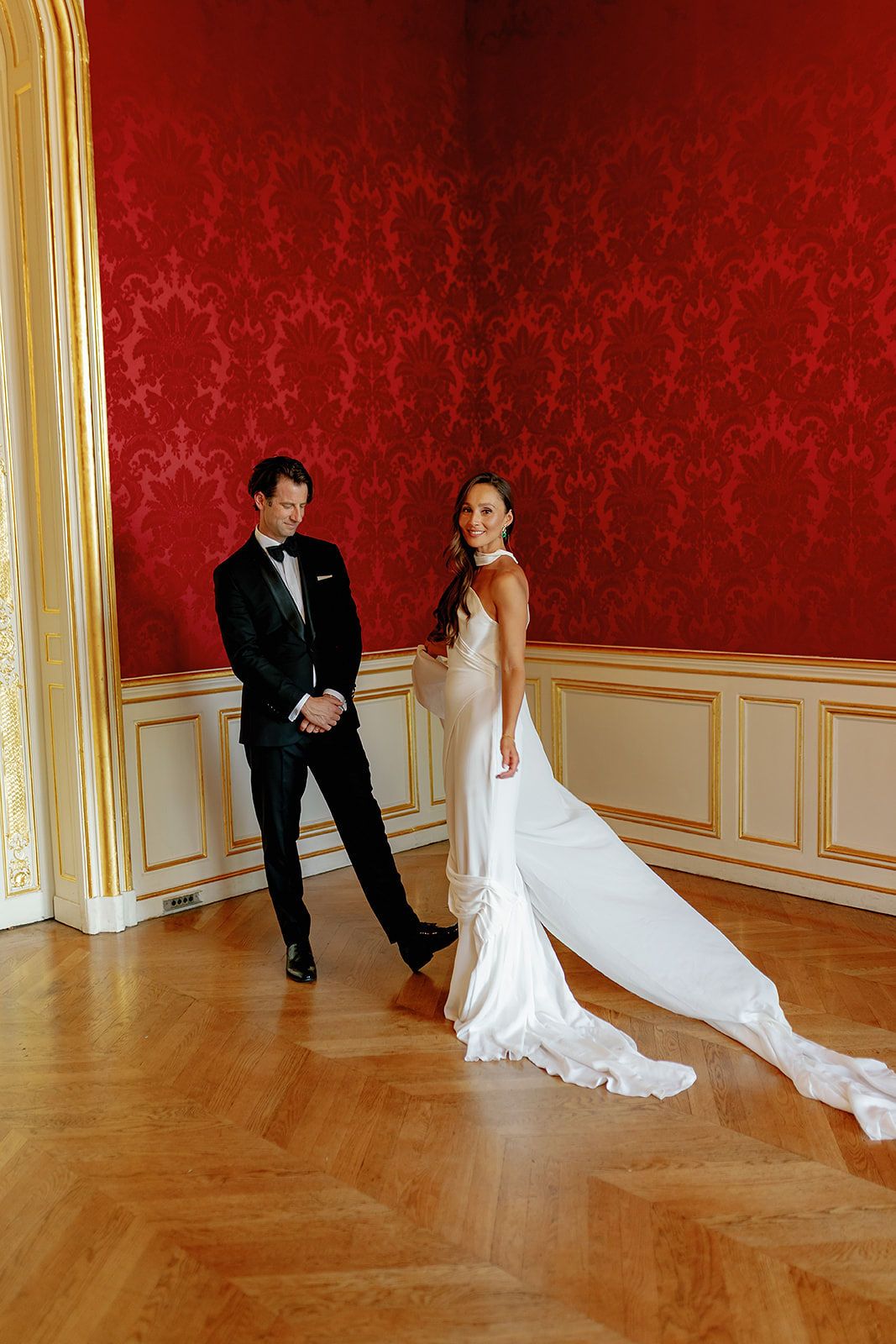 Elegant bride in white gown with train and groom in tuxedo posing in ornate red damask ballroom.