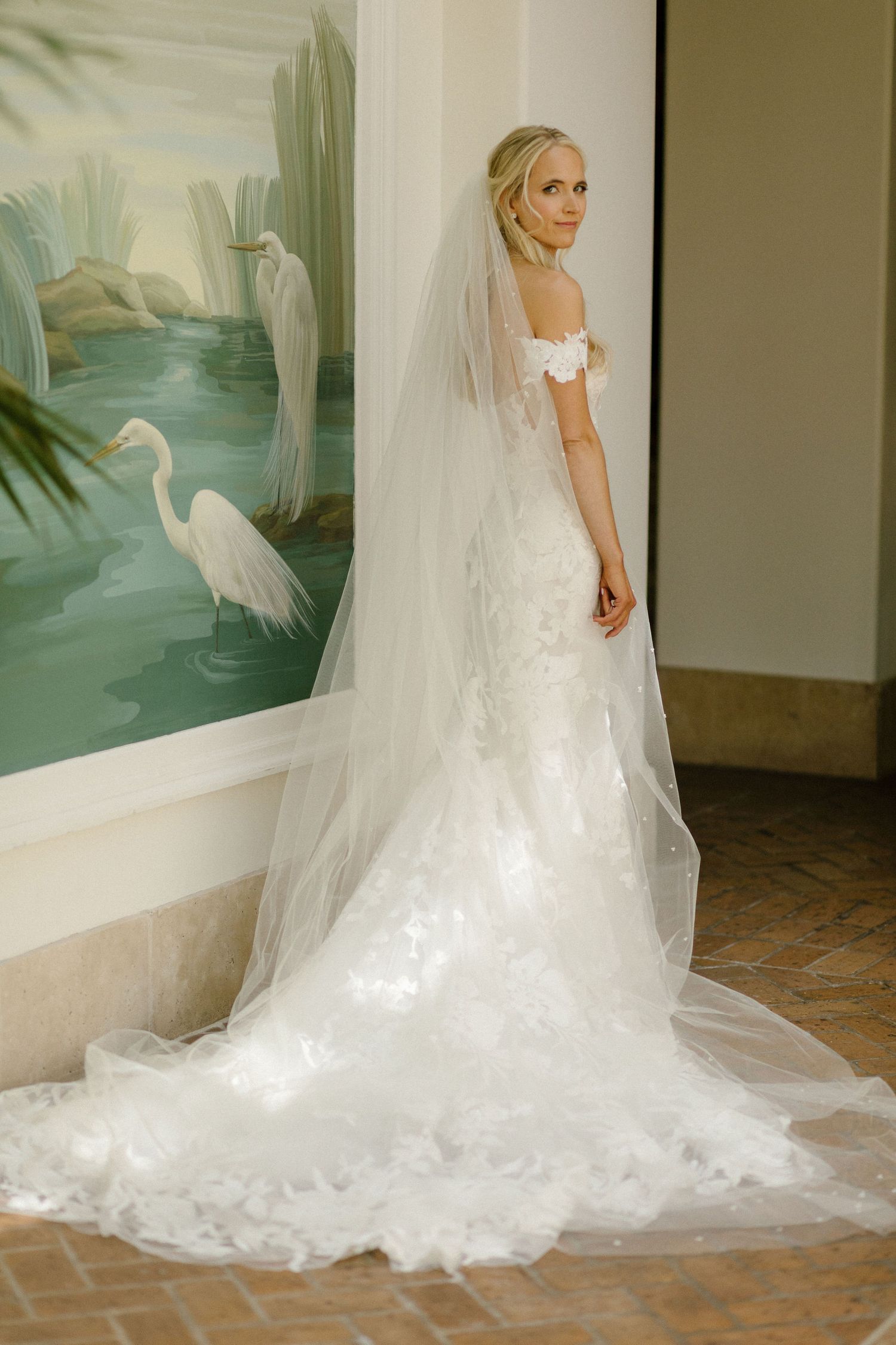 Bride in off-shoulder lace mermaid wedding gown with cathedral veil poses before egret painting.