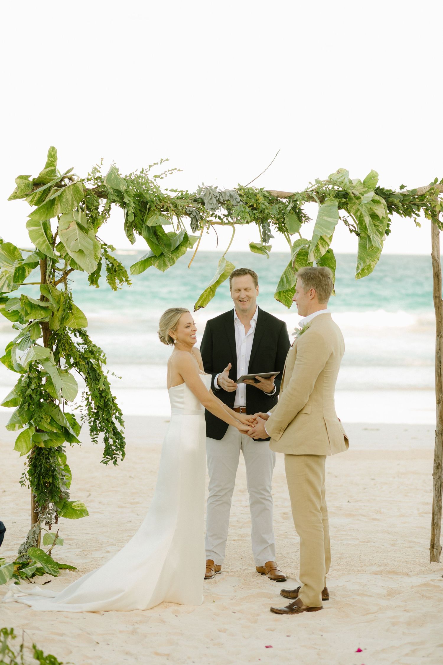 A beachside wedding ceremony with a couple under a lush green floral arch near the turquoise ocean.