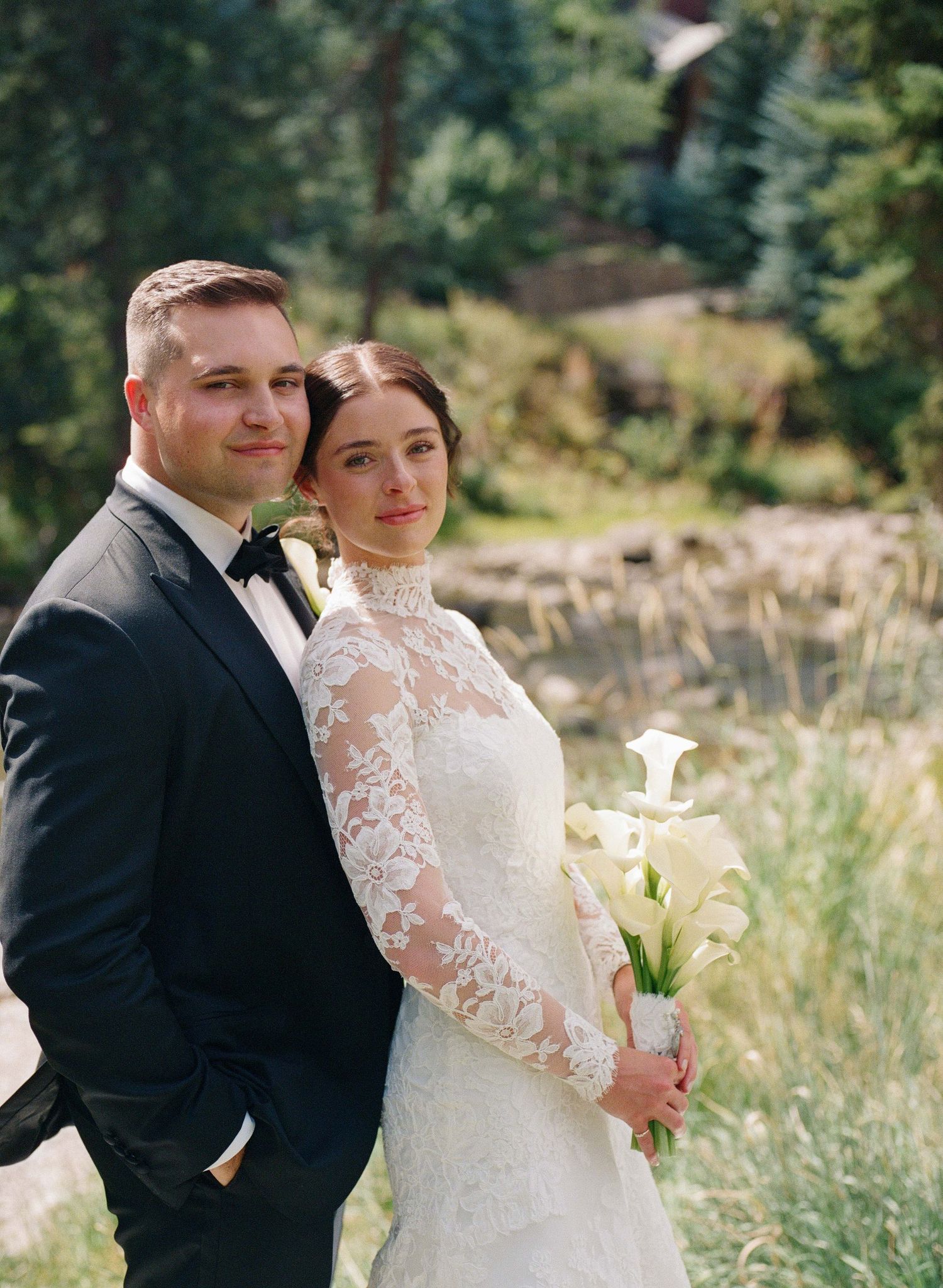 Bride in lace long-sleeve gown holding calla lilies stands with groom in black tuxedo outdoors in a forest setting.