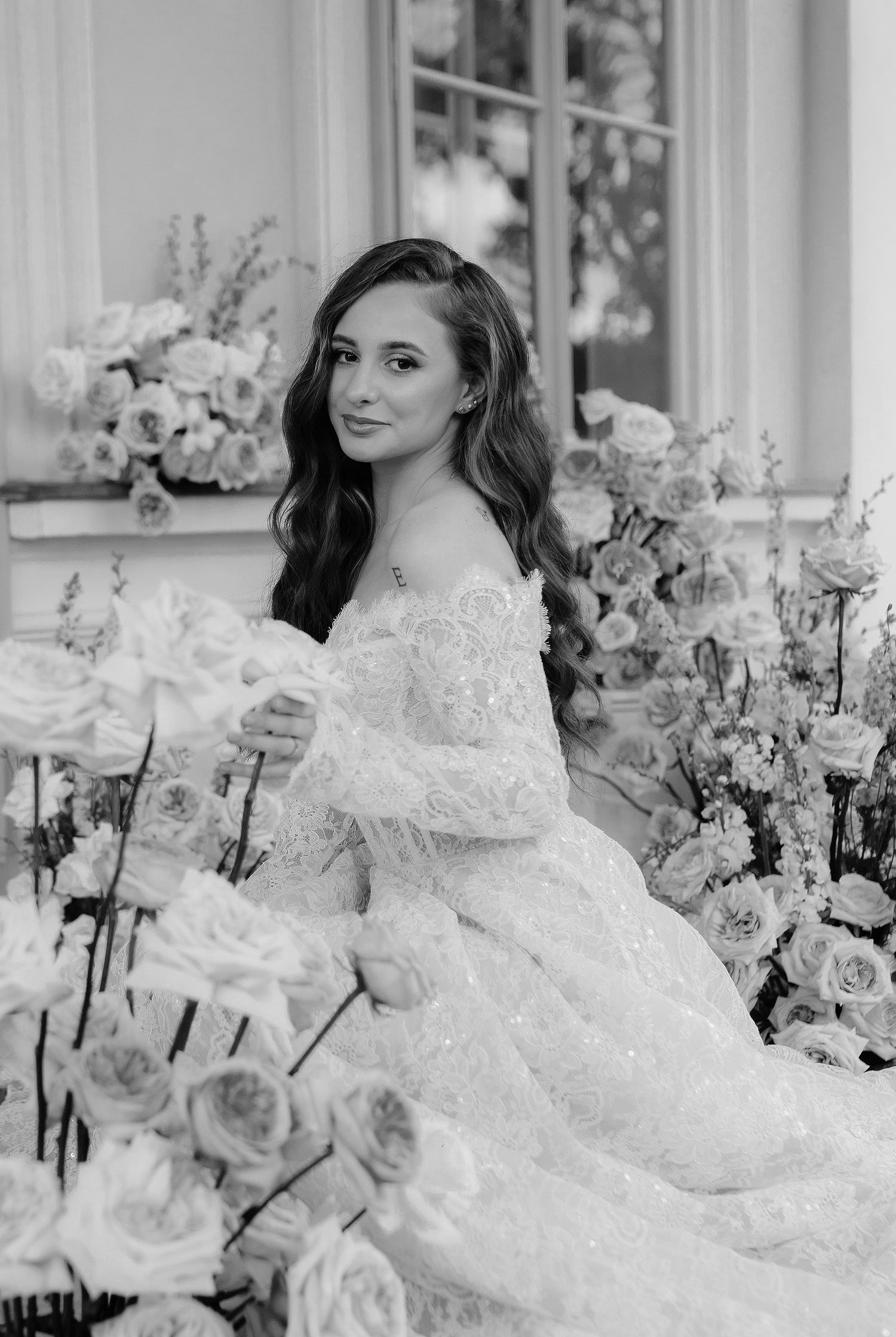 Black and white photograph of an elegant bride in a lace wedding dress surrounded by beautiful floral arrangements.