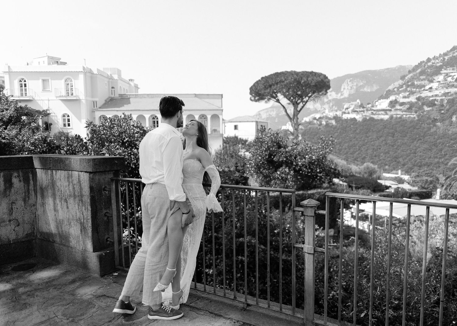 A black and white photograph overlooking an Italian coastal town with a couple embracing on a terrace.