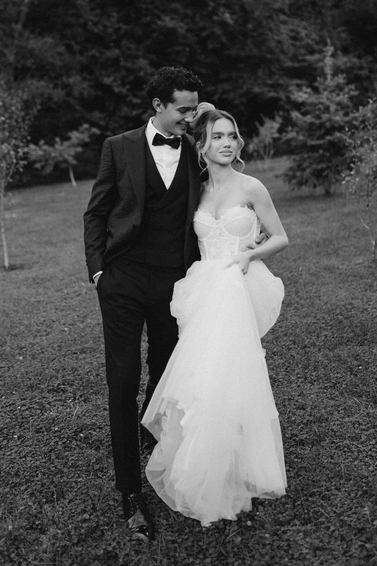 A black and white wedding photo showing a couple outdoors, with the bride in a strapless gown and groom in classic tuxedo.