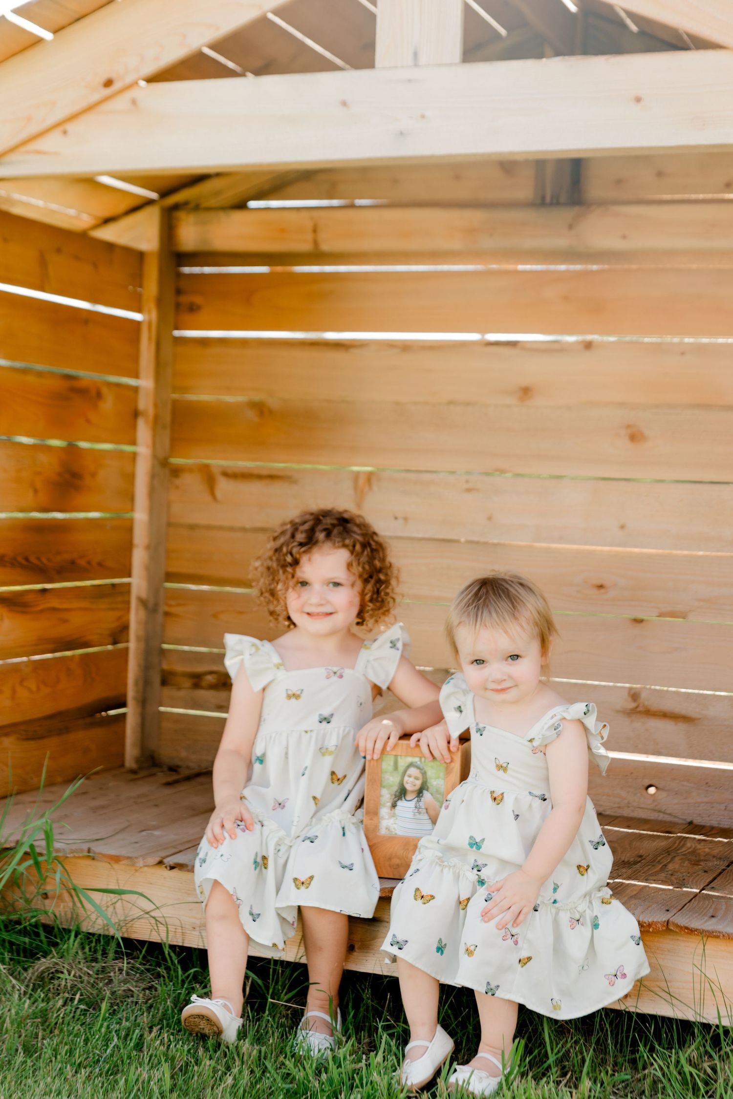 Two young children in matching butterfly print dresses sit together on wooden steps outside.