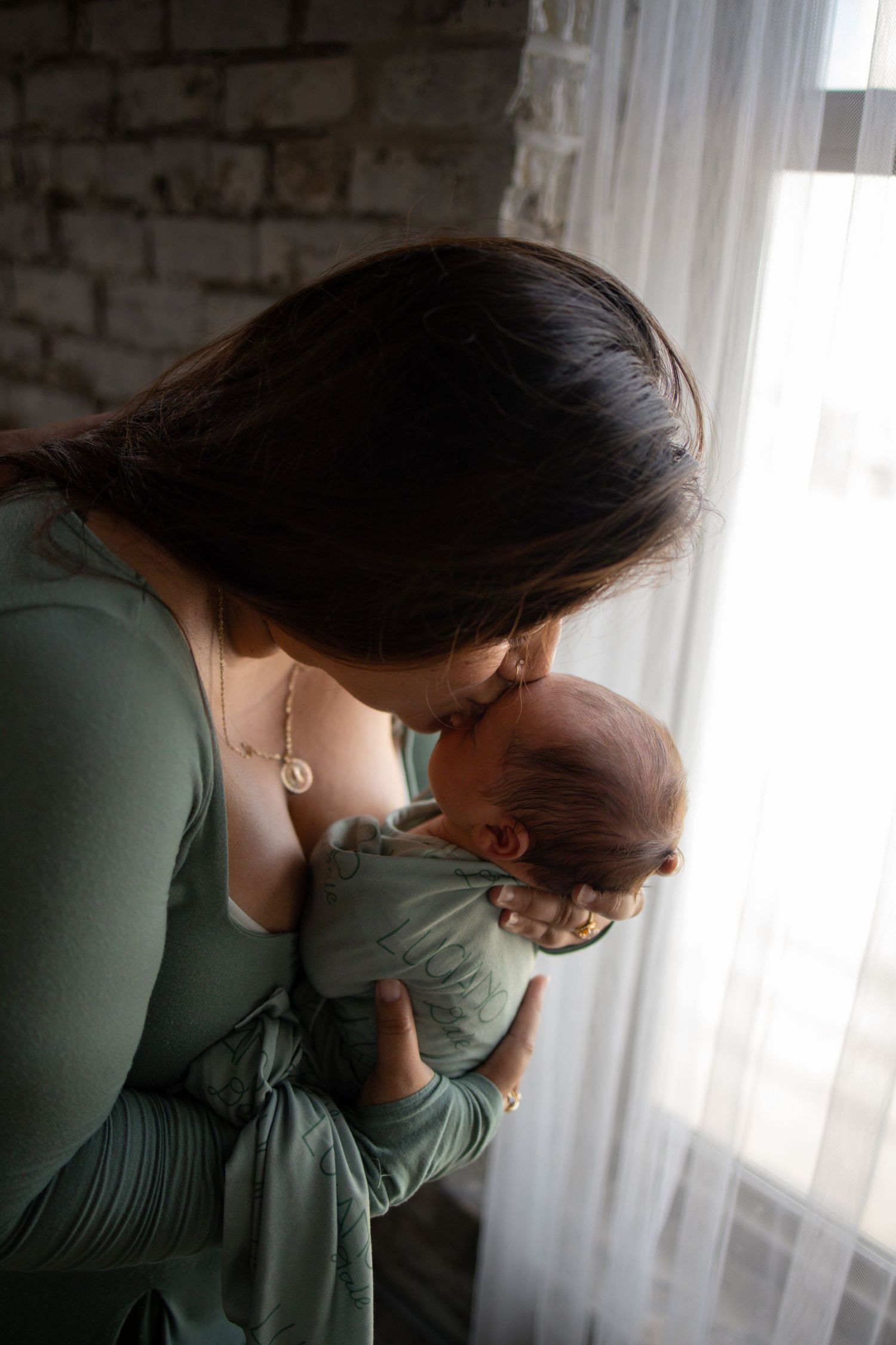 A tender moment between parent and newborn baby against a brick wall and sheer curtain background.