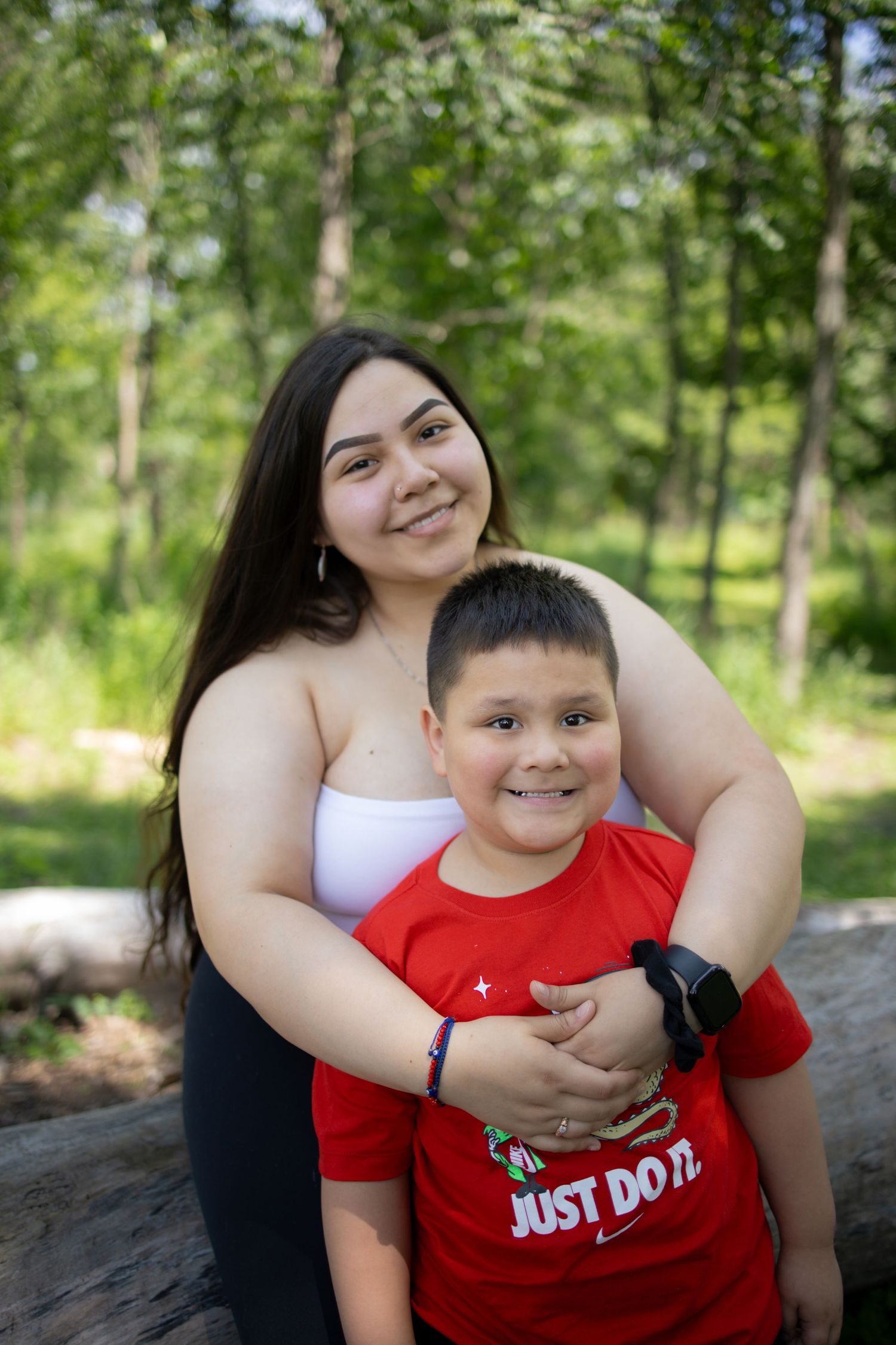 A warm embrace outdoors against a lush forest background, wearing casual summer attire including a red Nike shirt.