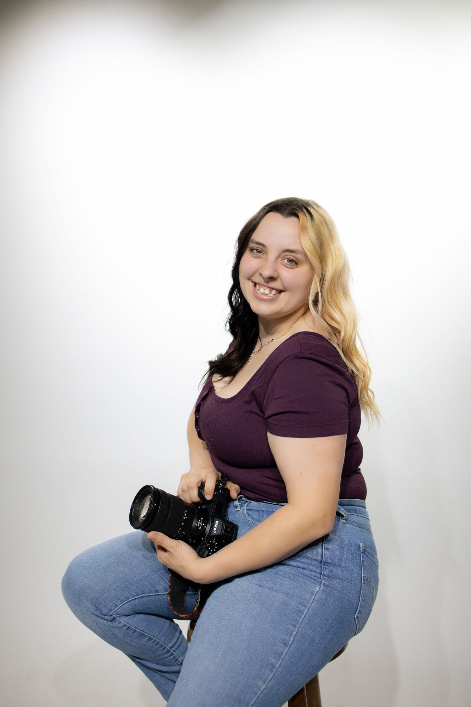 Photographer wearing burgundy top and jeans sits holding professional camera against white background.
