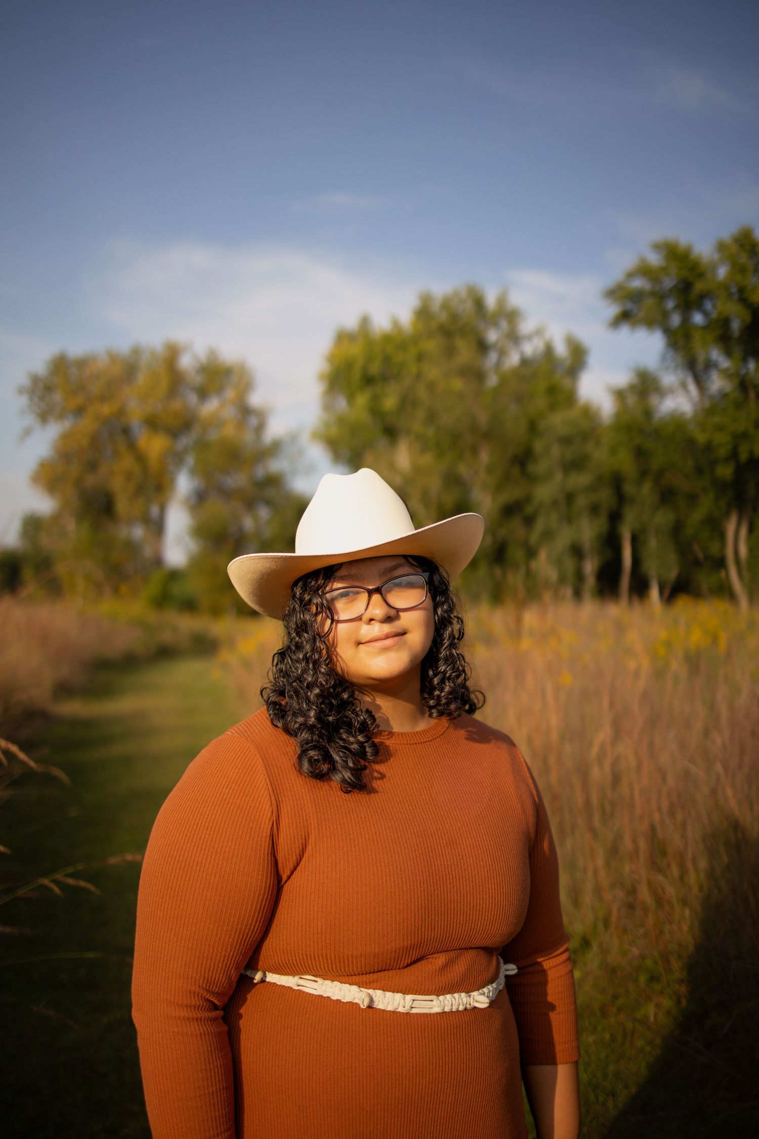 Person in a brown sweater and cowboy hat poses in a sunlit autumn field with tall grass and trees in the background.