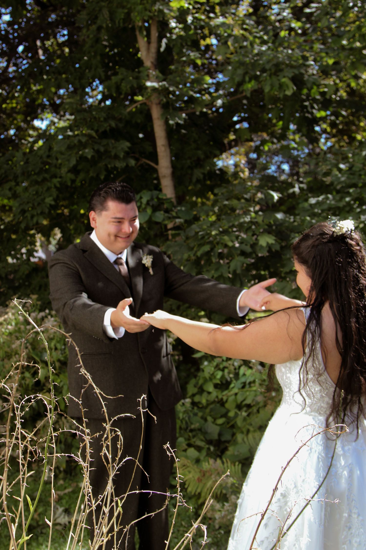 Couple in wedding attire playfully dance and twirl together in a lush green outdoor setting.