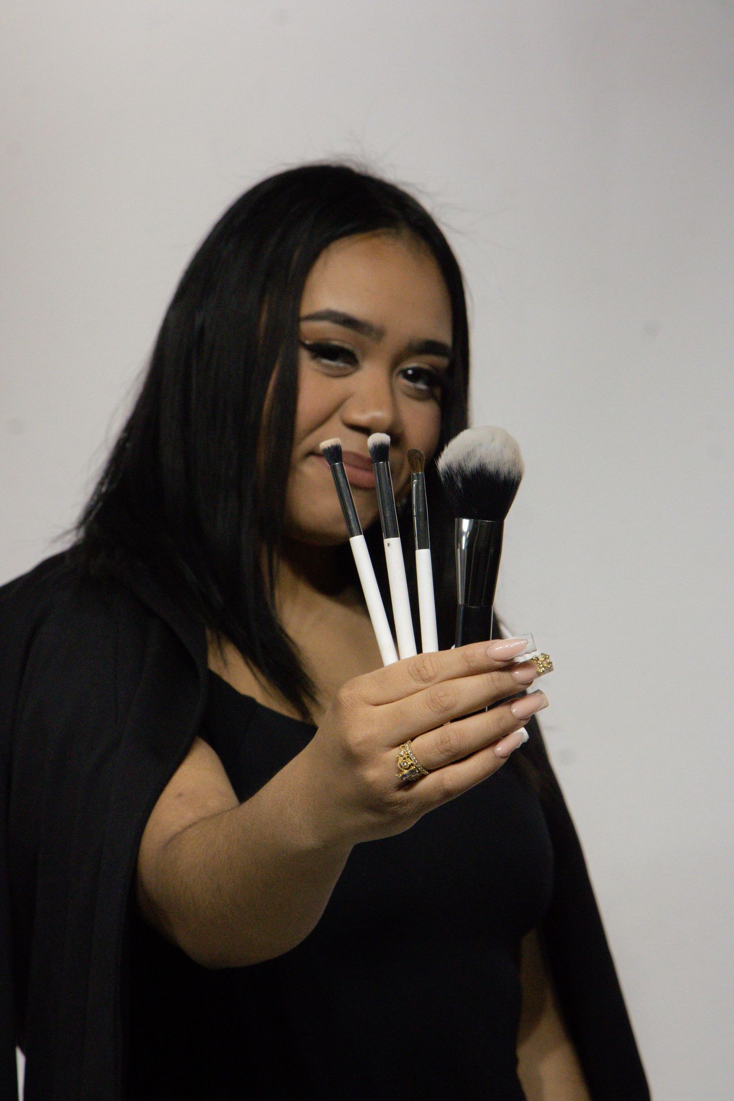 A makeup artist smiles while holding up a collection of professional makeup brushes against a gray background.
