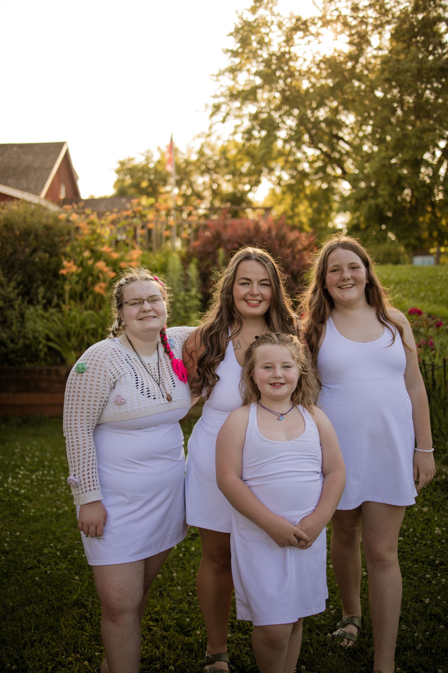 Four people wearing white dresses pose together in a garden during golden hour with trees and flowers in the background.