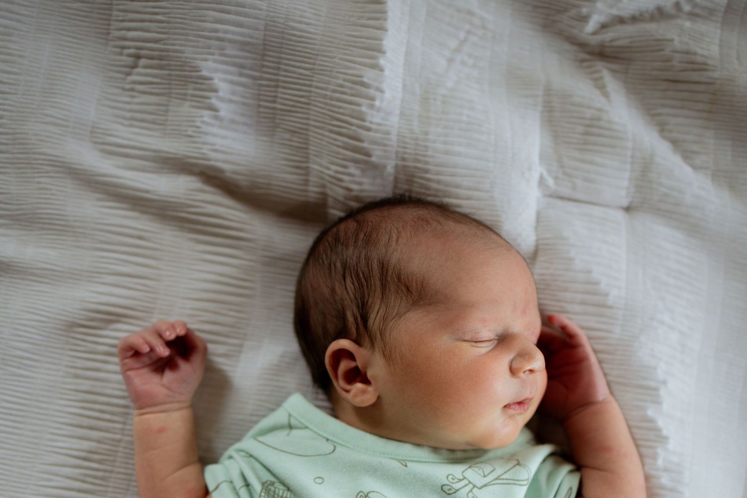 A newborn baby sleeps peacefully in white bedding while wearing a light mint green onesie.