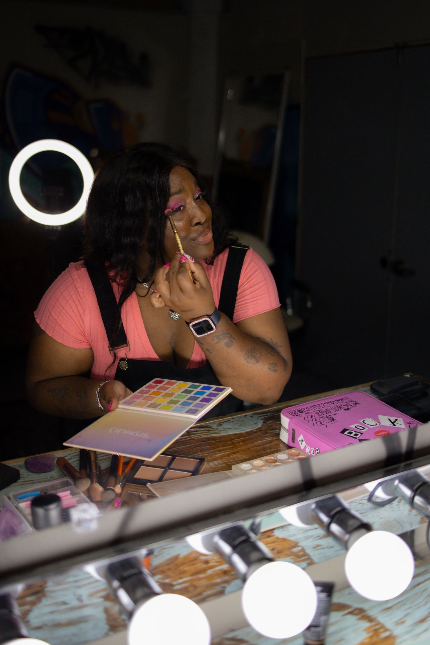 Someone in a pink top applies makeup at a well-lit vanity table with beauty products and a color palette visible.