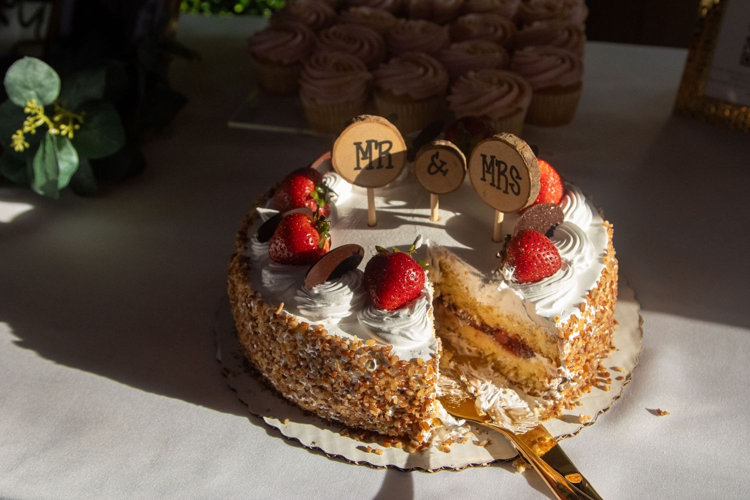 A sliced circular cream cake decorated with strawberries and chocolate pieces sitting on a white surface in soft lighting.