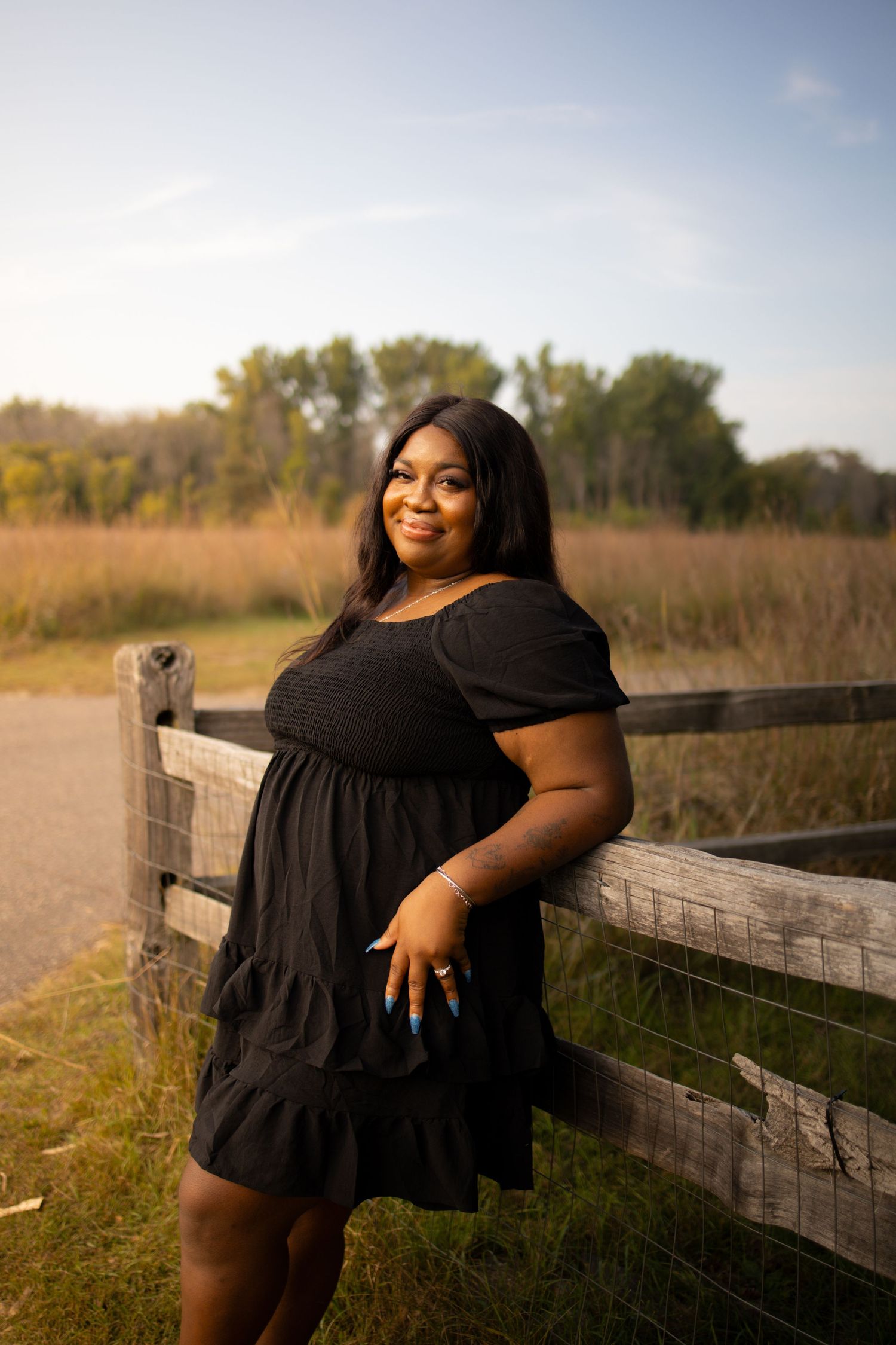 Woman in black dress leans on wooden fence in rural setting with autumn trees in background.