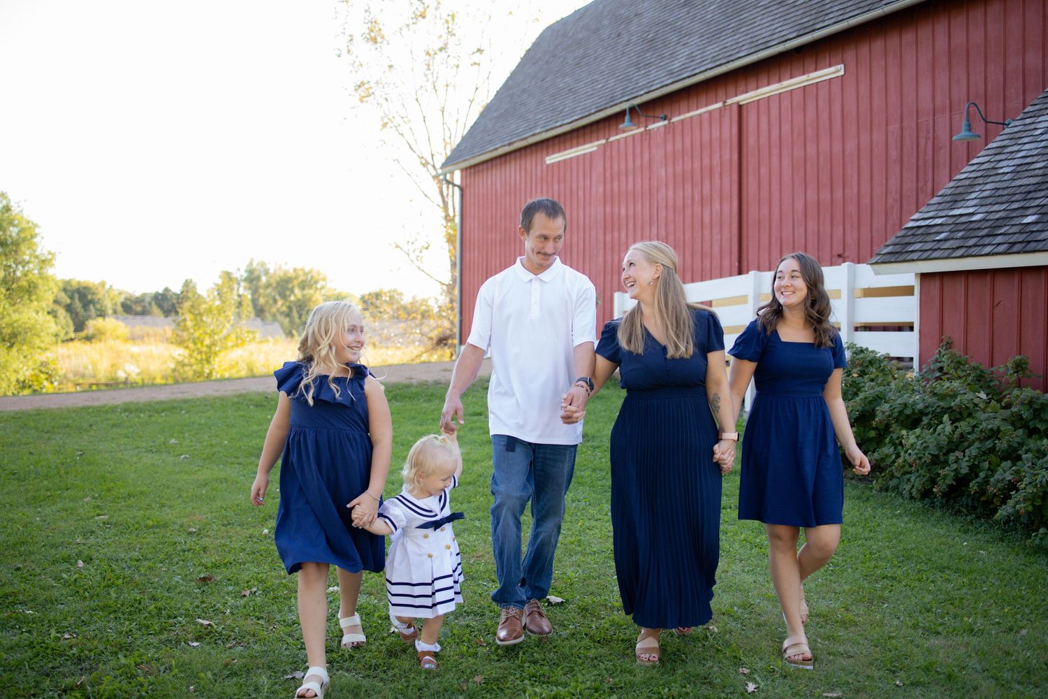 A family wearing navy blue and white outfits walks together near a red barn during a sunny outdoor photo session.