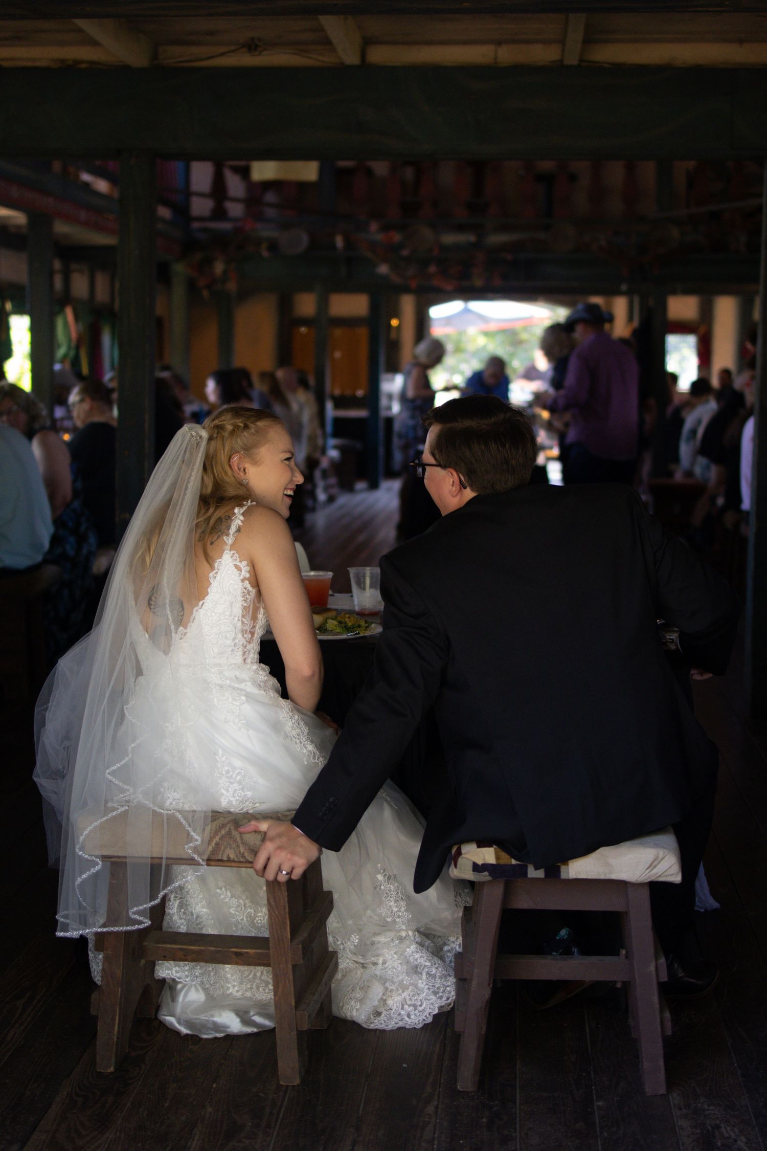A bride in a white lace wedding dress sits at a rustic wooden table during an indoor reception event.