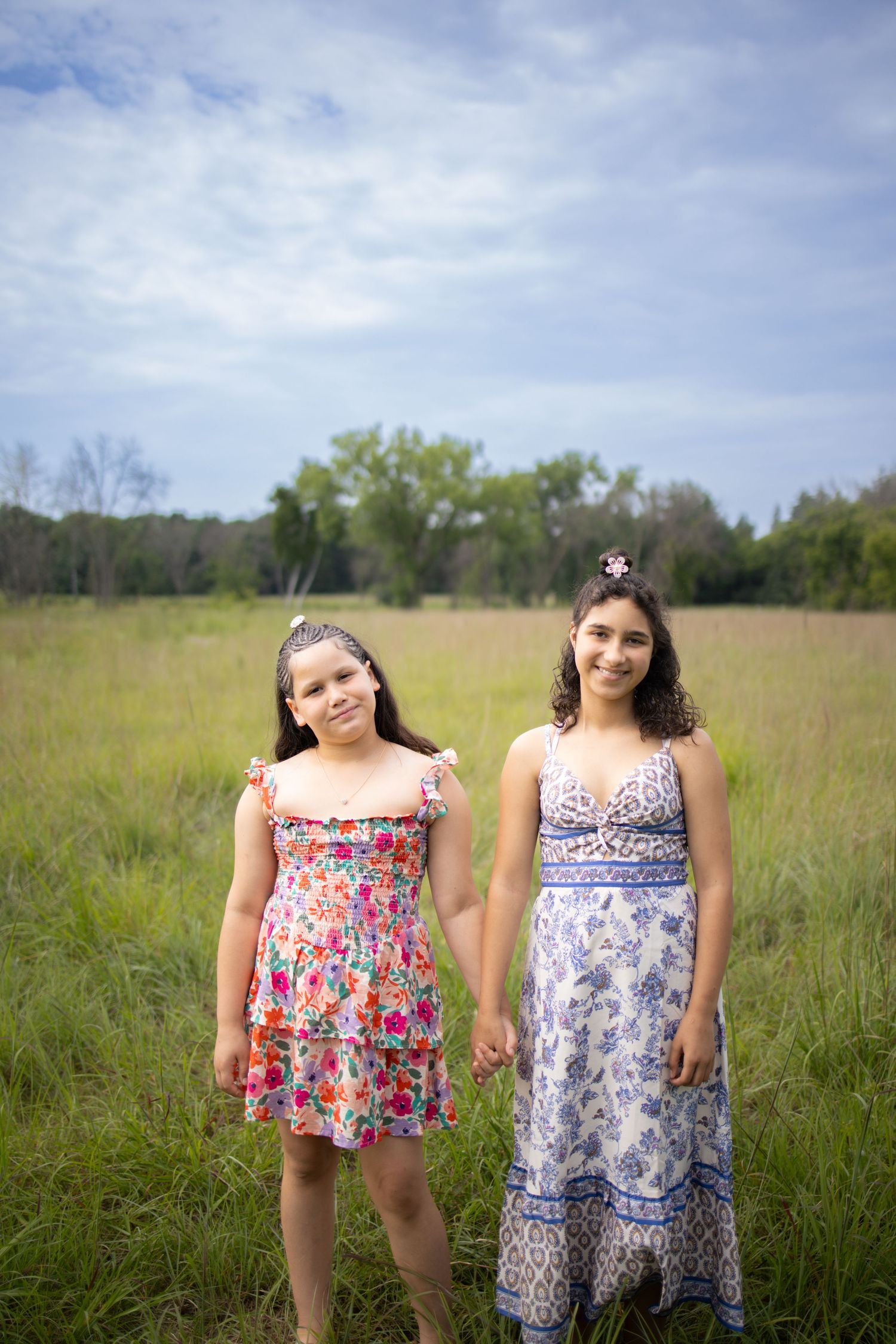 Two people wearing floral sundresses stand in a sunny meadow with tall grass and trees in the background.