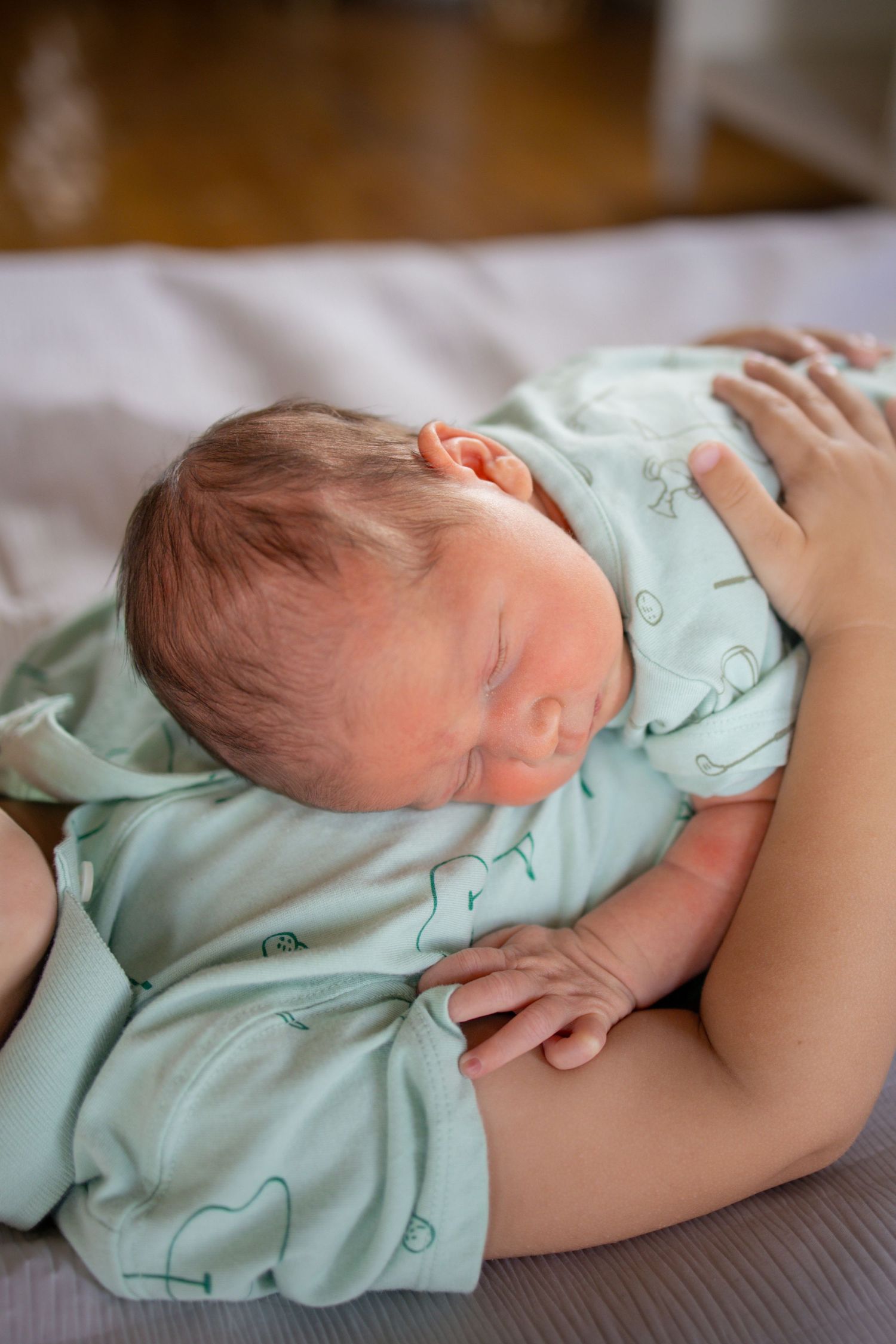 A newborn baby wearing a mint green onesie sleeps peacefully on a dark brown surface.