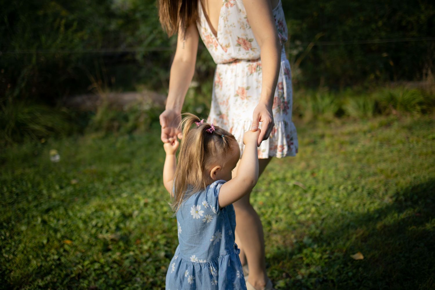 Person in denim overalls and white shirt walking through lush green garden path in summer light.