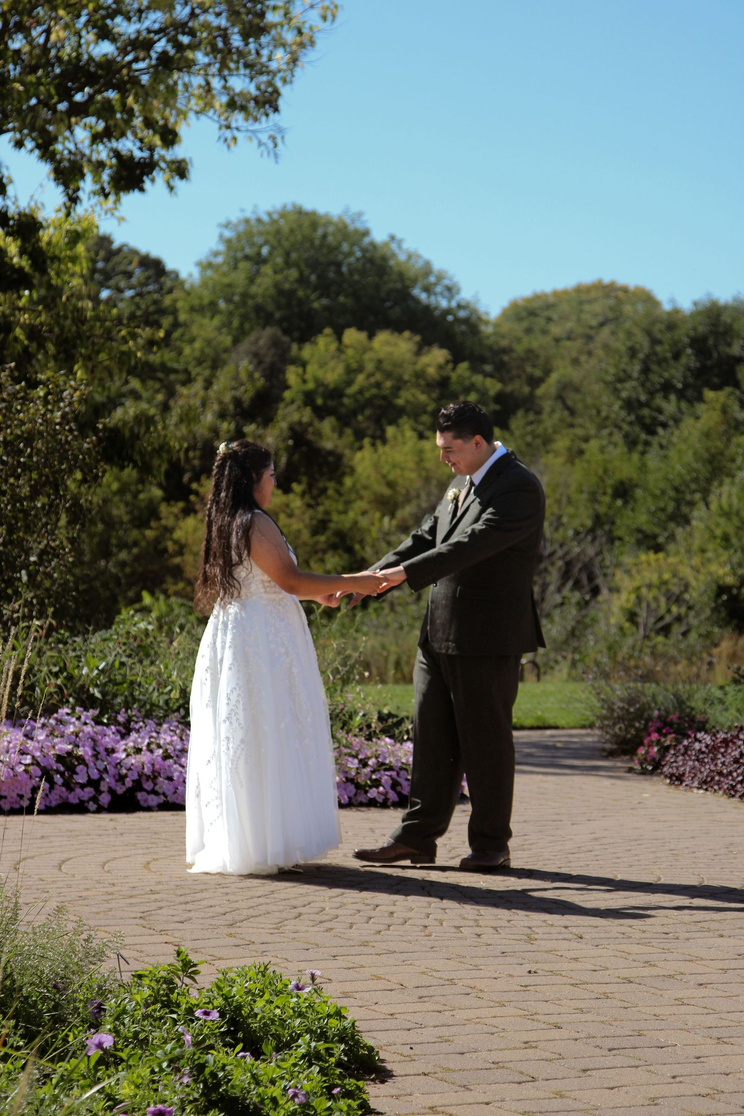 A couple holds hands during an outdoor wedding ceremony on a brick path surrounded by purple flowers and greenery.