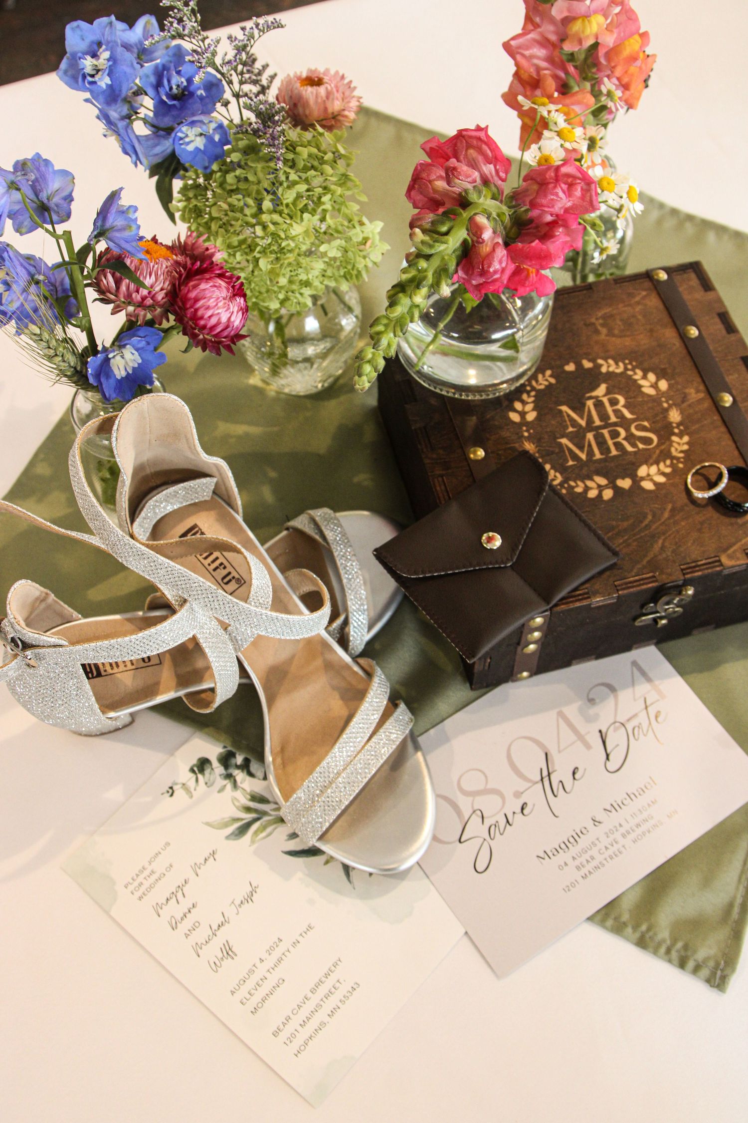 Wedding details including silver heels, floral arrangements in glass vases, save the date cards, and a Mr & Mrs wooden box.