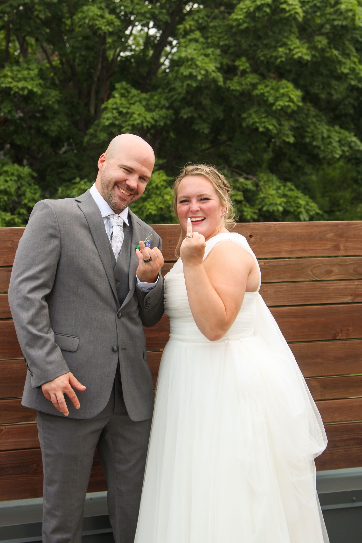 A couple in formal wedding attire posing playfully in front of a wooden wall outdoors.